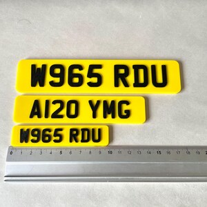 May include: Three yellow license plates with black lettering. The top plate reads "W965 RDU", the middle plate reads "AI20 YMG", and the bottom plate reads "W965 RDU". A ruler is placed below the plates, showing measurements in inches.