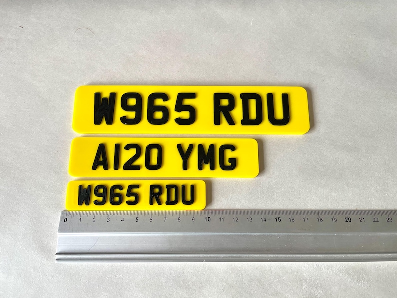 May include: Three yellow license plates with black lettering. The top plate reads "W965 RDU", the middle plate reads "AI20 YMG", and the bottom plate reads "W965 RDU". A ruler is placed below the plates, showing measurements in inches.