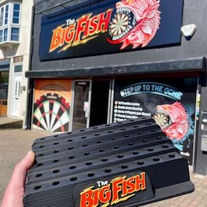 May include: Black plastic dart holder with multiple slots and the text "The Big Fish" in red and yellow. The holder is in front of a building with a sign that says "The Big Fish" and a dartboard graphic.