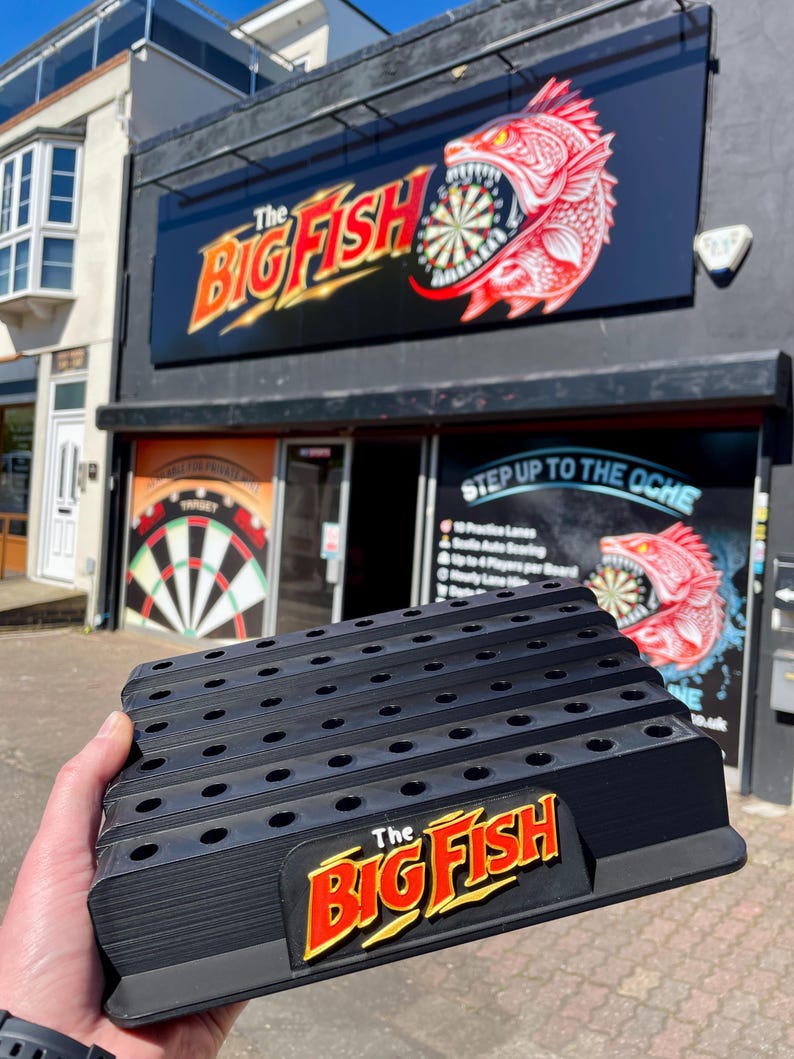 May include: Black plastic dart holder with multiple slots and the text "The Big Fish" in red and yellow. The holder is in front of a building with a sign that says "The Big Fish" and a dartboard graphic.