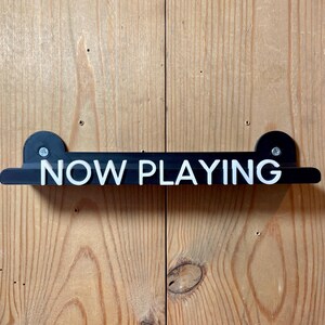 May include: Black wooden sign with white lettering that reads "NOW PLAYING" mounted on a wood wall.