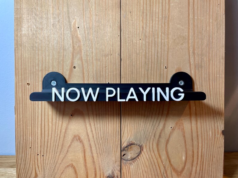 May include: Black wooden sign with white lettering that reads "NOW PLAYING" mounted on a wood wall.