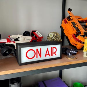 May include: A shelf displays a collection of items, including a white and red race car model, a gold trophy, and a large orange Lego car. A black sign with the words "ON AIR" in red is also present.