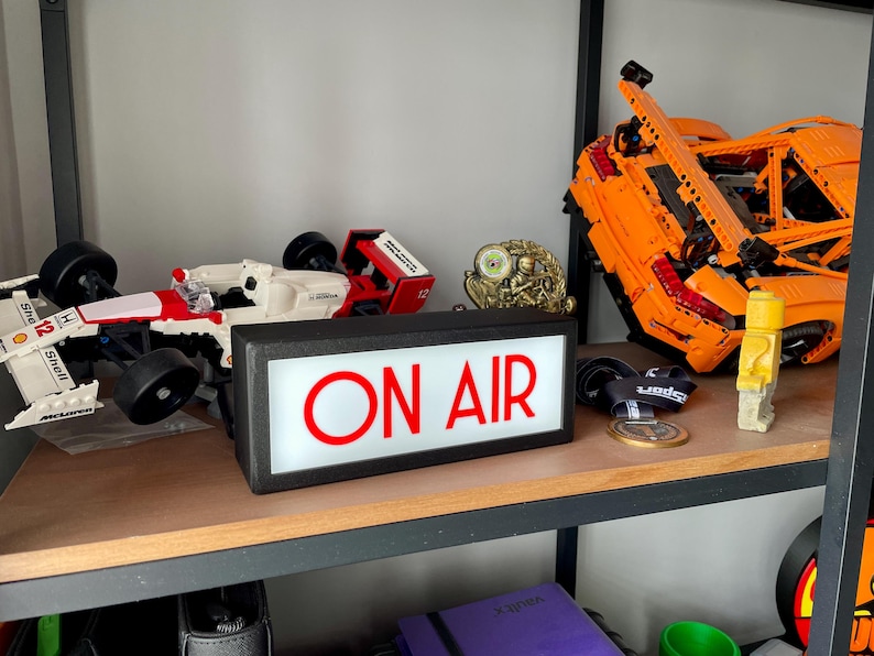 May include: A shelf displays a collection of items, including a white and red race car model, a gold trophy, and a large orange Lego car. A black sign with the words "ON AIR" in red is also present.
