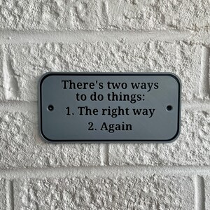 May include: A rectangular, gray metal sign with rounded corners is mounted on a white brick wall. The sign reads, "There's two ways to do things: 1. The right way 2. Again" in black text. Two small black dots are on either side.
