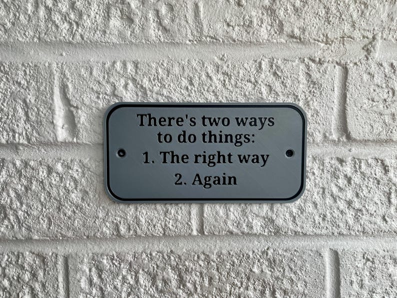 May include: A rectangular, gray metal sign with rounded corners is mounted on a white brick wall. The sign reads, "There's two ways to do things: 1. The right way 2. Again" in black text. Two small black dots are on either side.