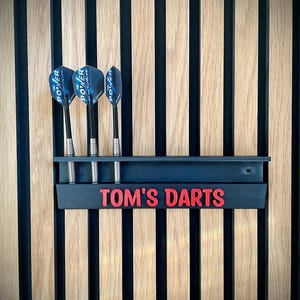May include: A black dart holder with the text "TOM'S DARTS" in red, mounted on a wooden wall. Three darts with blue and black flights are in the holder. The wall has vertical wood panels.