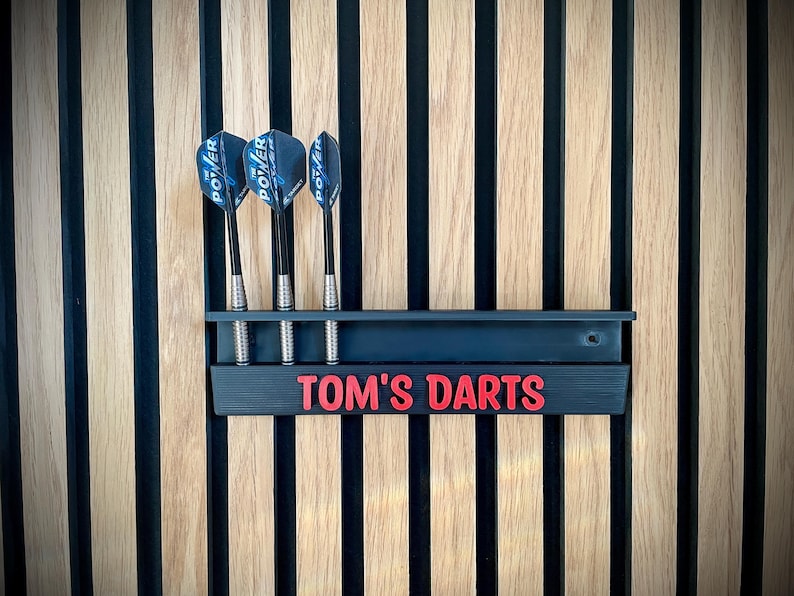 May include: A black dart holder with the text "TOM'S DARTS" in red, mounted on a wooden wall. Three darts with blue and black flights are in the holder. The wall has vertical wood panels.