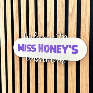 K&ouml;nnte beinhalten: Ein Klassenzimmerschild mit dem Text "Willkommen in Miss Honeys Klassenzimmer" in wei&szlig;er Schrift und lila Blockbuchstaben. Das Schild ist an einer Wand mit Holzlatten befestigt.