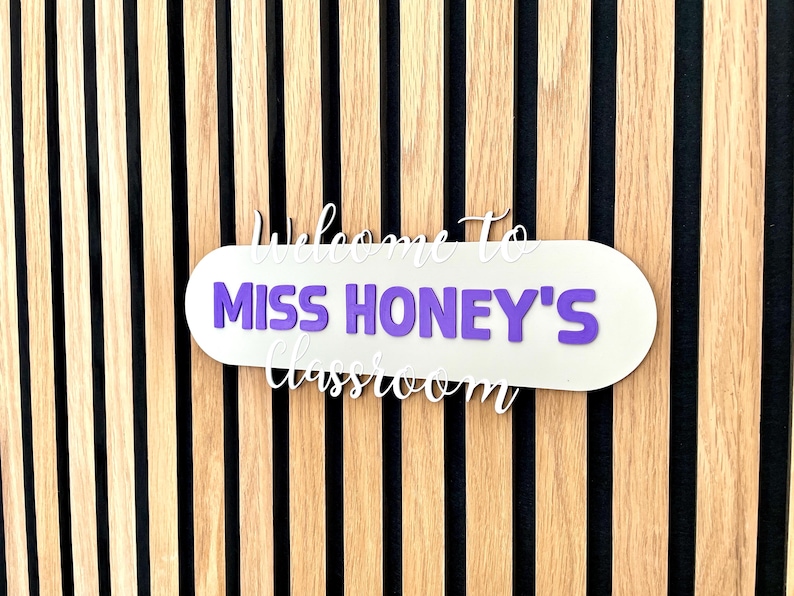 K&ouml;nnte beinhalten: Ein Klassenzimmerschild mit dem Text "Willkommen in Miss Honeys Klassenzimmer" in wei&szlig;er Schrift und lila Blockbuchstaben. Das Schild ist an einer Wand mit Holzlatten befestigt.