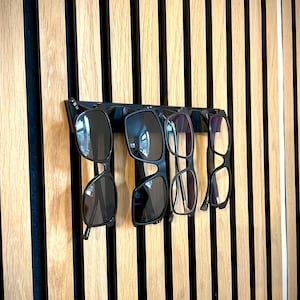 May include: A black eyewear holder with four pairs of glasses and sunglasses. The holder is mounted on a wooden wall with vertical stripes of light and dark wood. The glasses include two pairs of sunglasses and two pairs of eyeglasses.