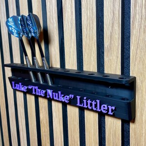 Puede incluir: Soporte negro para dardos con tres dardos. Los dardos tienen ca&ntilde;as negras y plumas Power azules y negras. El soporte tiene el nombre "Luke "The Nuke" Littler" en letras moradas. Montado en una pared de madera.
