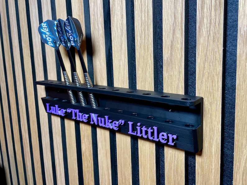 Puede incluir: Soporte negro para dardos con tres dardos. Los dardos tienen ca&ntilde;as negras y plumas Power azules y negras. El soporte tiene el nombre "Luke "The Nuke" Littler" en letras moradas. Montado en una pared de madera.
