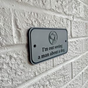 May include: A rectangular gray sign with a black border is mounted on a white brick wall. The sign features a dog silhouette and the phrase "I'm out seeing a man about a dog."
