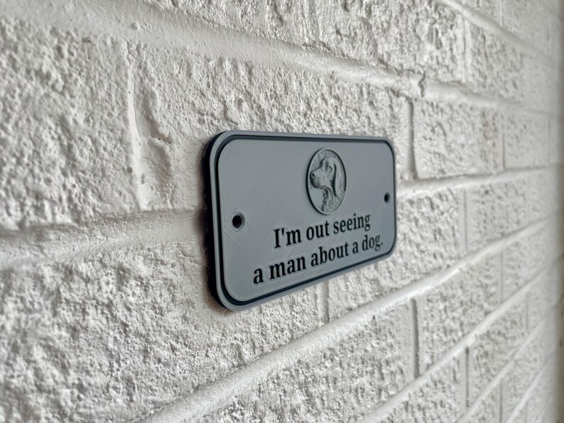 May include: A rectangular gray sign with a black border is mounted on a white brick wall. The sign features a dog silhouette and the phrase "I'm out seeing a man about a dog."