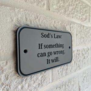 May include: A rectangular sign with rounded corners, mounted on a textured white brick wall. The sign is gray with a black border and features the text "Sod's Law: If something can go wrong, It will."