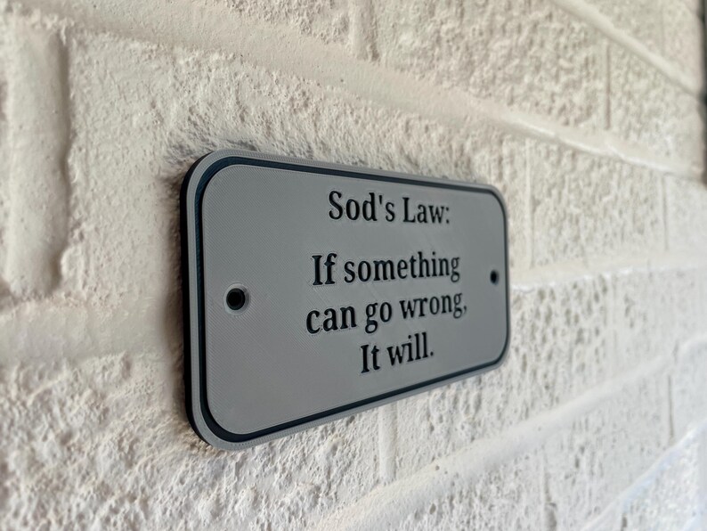 May include: A rectangular sign with rounded corners, mounted on a textured white brick wall. The sign is gray with a black border and features the text "Sod's Law: If something can go wrong, It will."