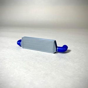 May include: A gray intercooler with horizontal fins and blue silicone couplers. This automotive part is designed to cool the air entering an engine, enhancing performance. The blue couplers connect to the intercooler's ends.
