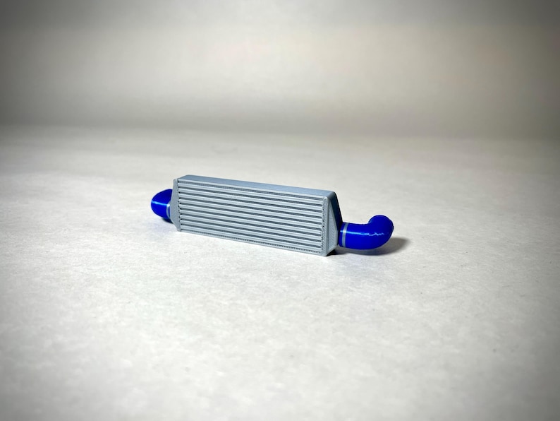 May include: A gray intercooler with horizontal fins and blue silicone couplers. This automotive part is designed to cool the air entering an engine, enhancing performance. The blue couplers connect to the intercooler's ends.