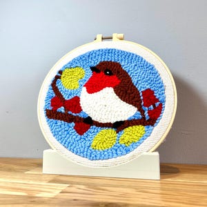 May include: A colorful needlepoint embroidery hoop with a robin perched on a branch. The robin is brown, red, and white, and the background is blue with yellow leaves.