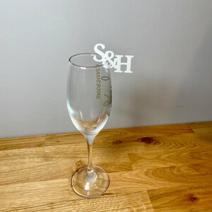 Champagne flute with a set of initial letters clipped onto the side. Drink topper is white and the photo shows a grey background.