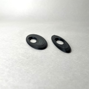 May include: Two black, oval-shaped objects with a hole in the center. The objects have a layered appearance, with each layer slightly offset from the one below. They are set against a plain, off-white background.