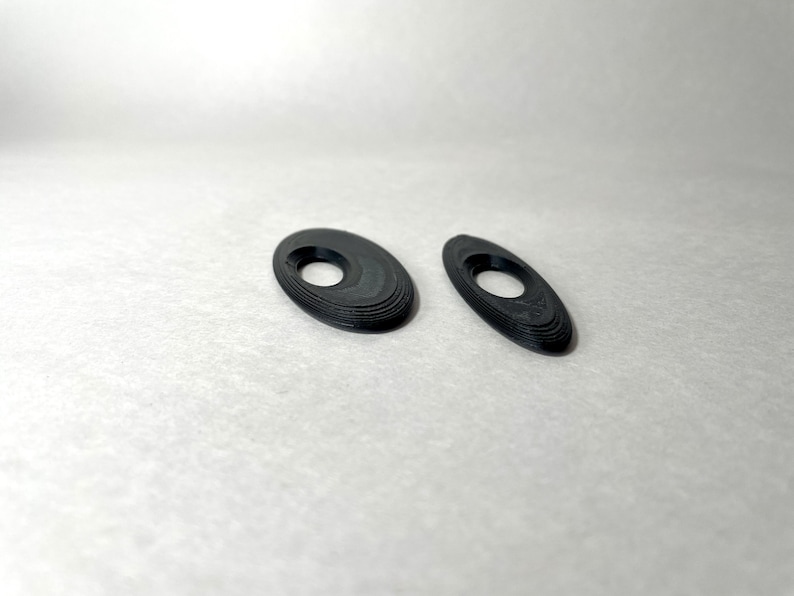 May include: Two black, oval-shaped objects with a hole in the center. The objects have a layered appearance, with each layer slightly offset from the one below. They are set against a plain, off-white background.