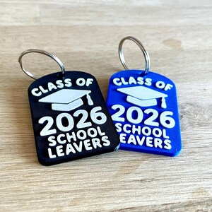 May include: Two keychains, one black and one blue, with silver key rings. Each keychain features the text "CLASS OF 2026 SCHOOL LEAVERS" in white, along with a graduation cap graphic. The keychains are on a wooden surface.