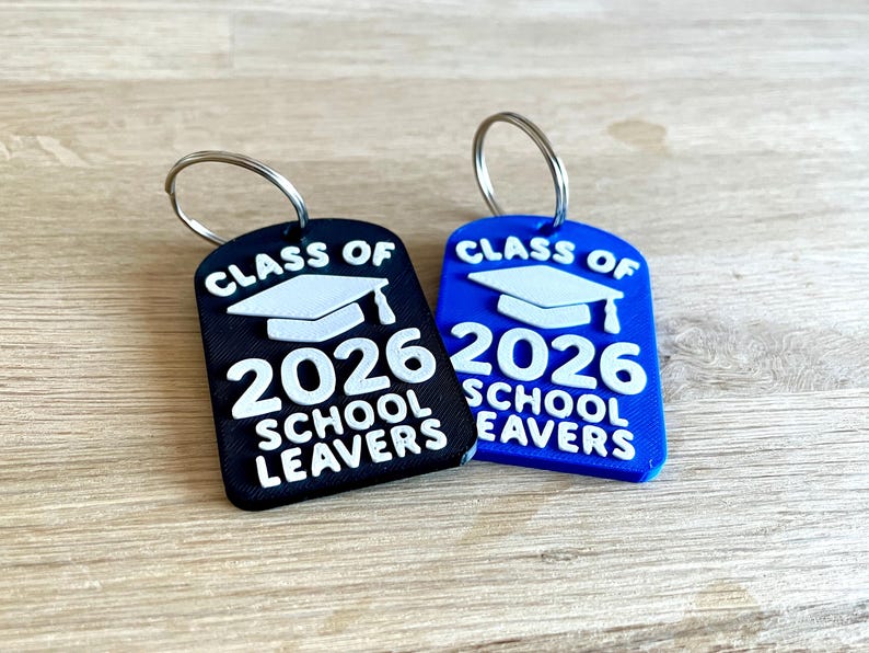 May include: Two keychains, one black and one blue, with silver key rings. Each keychain features the text "CLASS OF 2026 SCHOOL LEAVERS" in white, along with a graduation cap graphic. The keychains are on a wooden surface.