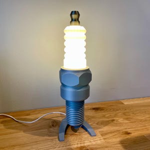 May include: A gray and white table lamp shaped like a spark plug with a white light bulb turned on. The lamp is sitting on a wooden table.