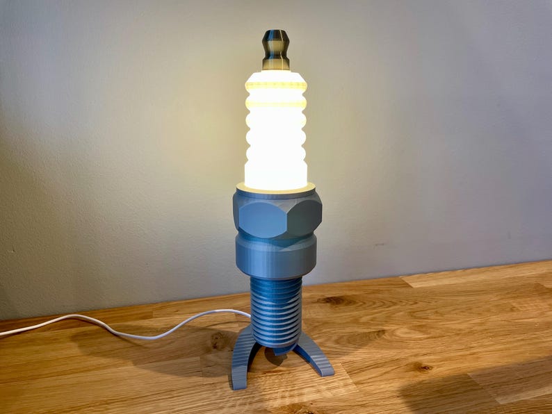 Oversized spark plug lamp with the white ceramic section designed as a light shade. Bronze top section, white shade and silver base. With a white cable. Stood on a wooden desk and grey background.
