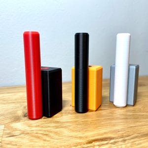 Op de afbeelding: Drie cilindrische voorwerpen, elk in een andere kleur, staan rechtop in kleine rechthoekige houders. De voorwerpen zijn rood, zwart en wit. De houders zijn zwart, oranje en grijs.