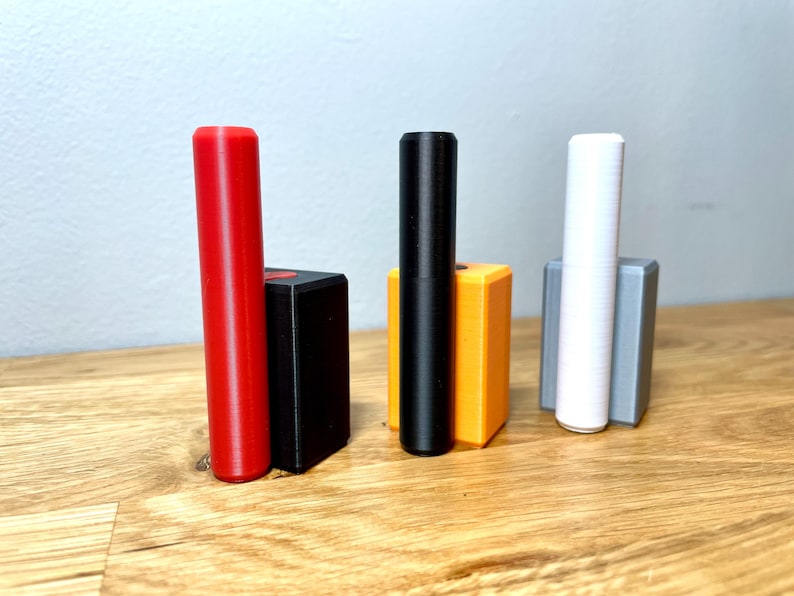 Op de afbeelding: Drie cilindrische voorwerpen, elk in een andere kleur, staan rechtop in kleine rechthoekige houders. De voorwerpen zijn rood, zwart en wit. De houders zijn zwart, oranje en grijs.