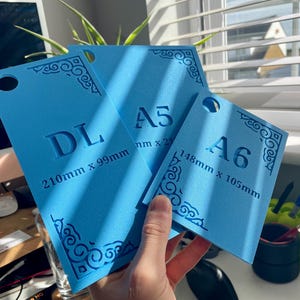 May include: Three bright blue rectangular templates with rounded corners and decorative corner patterns. The templates are labeled DL 210mm x 99mm, A5, and A6 148mm x 105mm. Each piece features a circular hole at the top for hanging or storage.