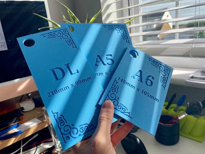 May include: Three bright blue rectangular templates with rounded corners and decorative corner patterns. The templates are labeled DL 210mm x 99mm, A5, and A6 148mm x 105mm. Each piece features a circular hole at the top for hanging or storage.