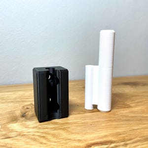 Op de afbeelding: Twee 3D-geprinte objecten, een zwart en een wit. Het zwarte object is een rechthoekig blok met drie gaten aan de voorkant. Het witte object is een hoge, dunne cilinder met een afgeronde bovenkant.