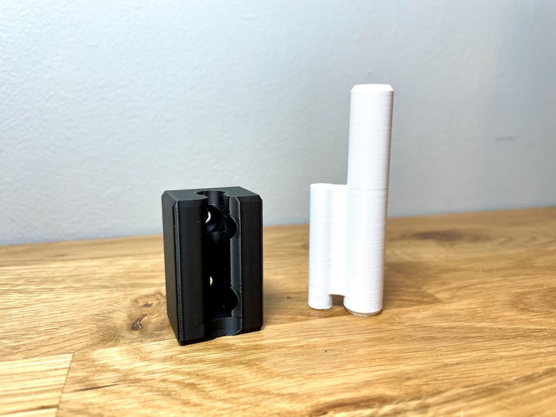 Op de afbeelding: Twee 3D-geprinte objecten, een zwart en een wit. Het zwarte object is een rechthoekig blok met drie gaten aan de voorkant. Het witte object is een hoge, dunne cilinder met een afgeronde bovenkant.