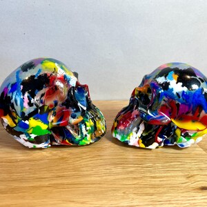 May include: Two colourful, hand-painted human skulls, each with a different abstract design. The skulls are made of resin and are approximately 15 cm tall.