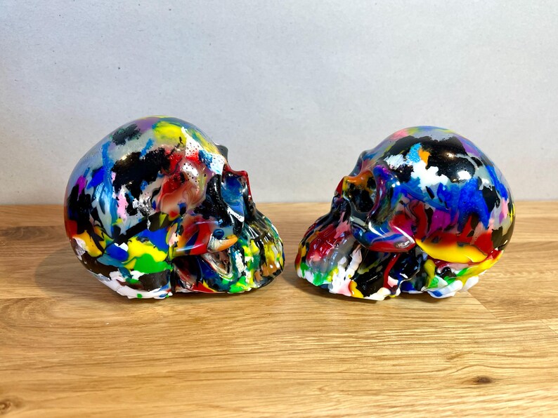 May include: Two colourful, hand-painted human skulls, each with a different abstract design. The skulls are made of resin and are approximately 15 cm tall.