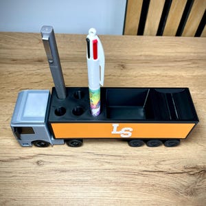 Pu&ograve; includere: Un organizer da scrivania a forma di camion grigio e arancione con ruote nere. L'organizer ha scomparti per penne e altri articoli per l'ufficio. Ci sono due penne nell'organizer. Il lato del camion ha le lettere "LS" in bianco.