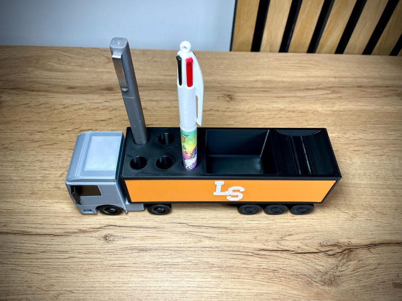Pu&ograve; includere: Un organizer da scrivania a forma di camion grigio e arancione con ruote nere. L'organizer ha scomparti per penne e altri articoli per l'ufficio. Ci sono due penne nell'organizer. Il lato del camion ha le lettere "LS" in bianco.