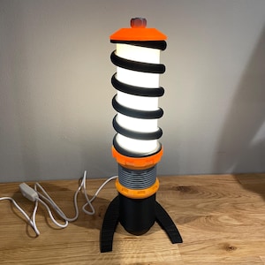 May include: A black and orange table lamp shaped like a car shock absorber with a white light bulb. The lamp has a black base with a rounded bottom and two feet.