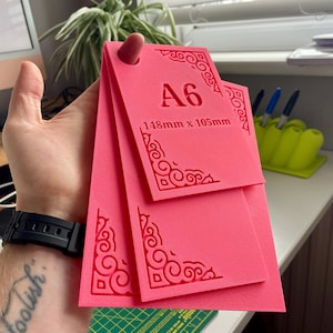May include: A set of three bright pink rectangular templates with rounded corners and intricate scrollwork patterns embossed on the edges. The top template features the text A6 and 148mm x 105mm. Each template has a circular hole at the top.
