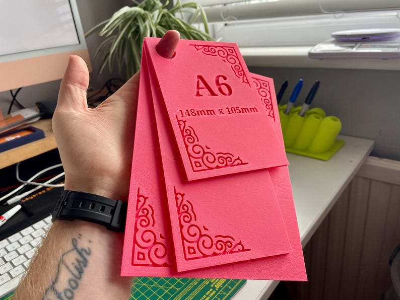May include: A set of three bright pink rectangular templates with rounded corners and intricate scrollwork patterns embossed on the edges. The top template features the text A6 and 148mm x 105mm. Each template has a circular hole at the top.