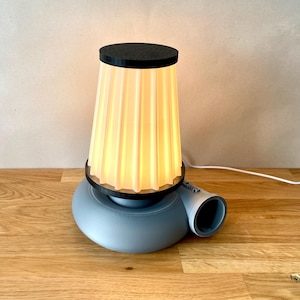 May include: A grey table lamp with a pleated white shade and a black top. The lamp has a unique base with a curved shape and a small opening on the side.