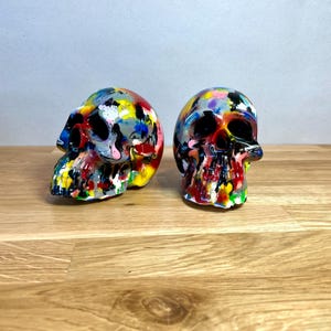 May include: Two colourful painted human skulls, one is facing left and the other is facing right. The skulls are painted in a variety of colours, including red, yellow, blue, green, and black.