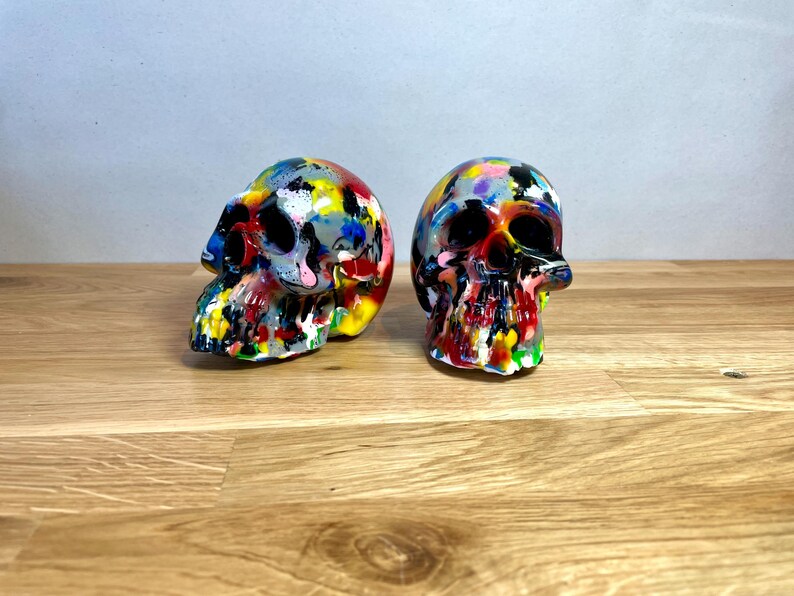 May include: Two colourful painted human skulls, one is facing left and the other is facing right. The skulls are painted in a variety of colours, including red, yellow, blue, green, and black.