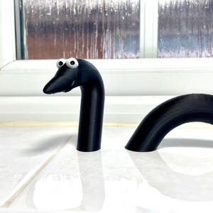 May include: A black, 3D printed, snake-like toy with googly eyes. The toy is in two pieces, with the head and neck separate from the body.