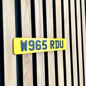 May include: A yellow rectangular license plate with the text "W965 RDU" in dark gray lettering. The plate is set against a backdrop of vertical wooden slats with alternating light and dark tones.