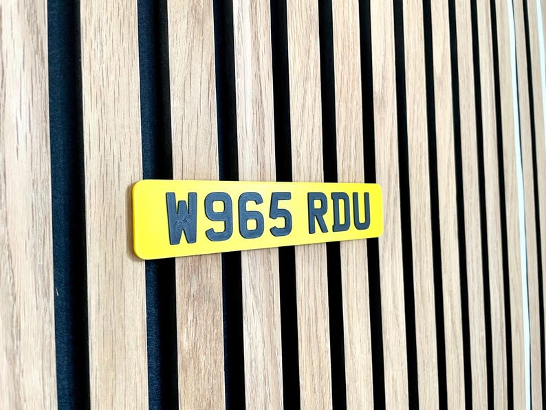 May include: A yellow rectangular license plate with the text "W965 RDU" in dark gray lettering. The plate is set against a backdrop of vertical wooden slats with alternating light and dark tones.